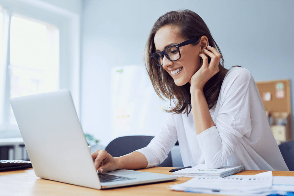 A woman wearing glasses smiles as she uses her laptop to work.