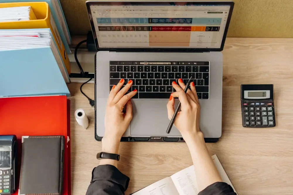 A woman's hands on a laptop, calculator, and office supplies, indicating work or productivity.