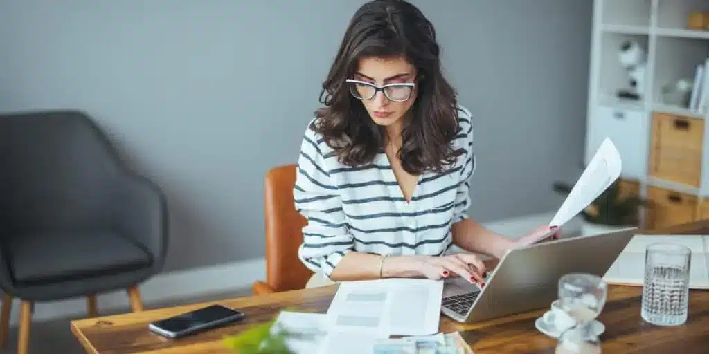 A woman sitting at a desk with a laptop and papers, working diligently in a home office setting.